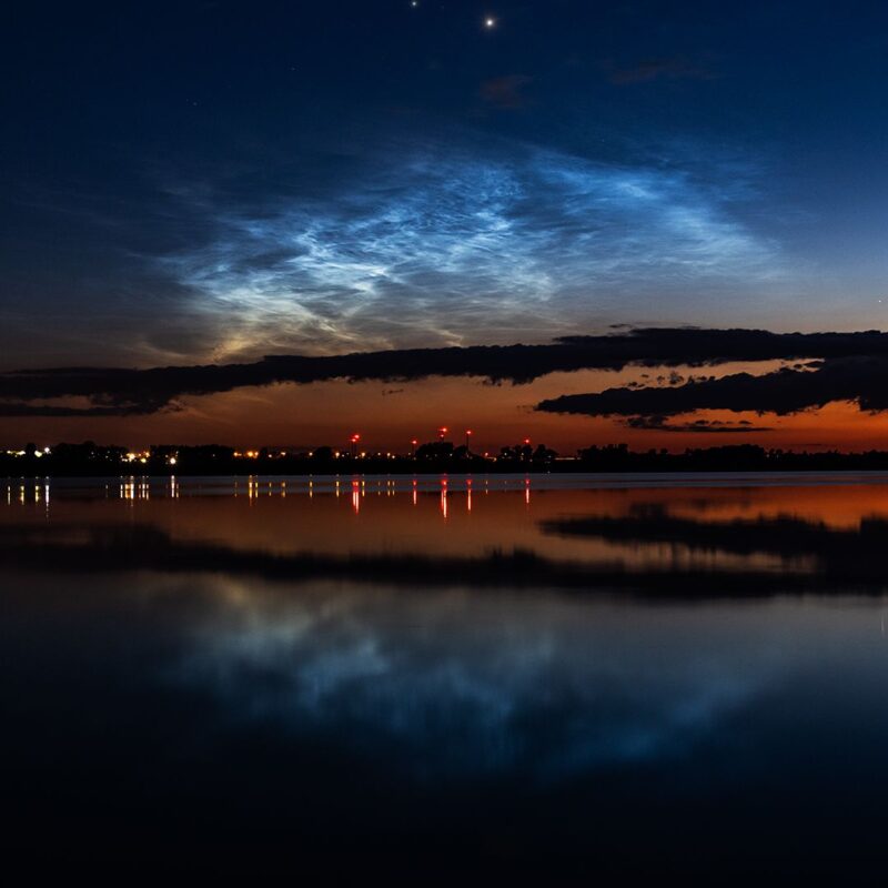 Glowing blue clouds in a dark sky reflected in a lake with town lights on opposite shore.