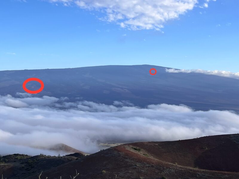 Mountain in the background and a mountain with a crater in the foreground. There are clouds between them. Two circles around 2 white dots in the bacground mountain.