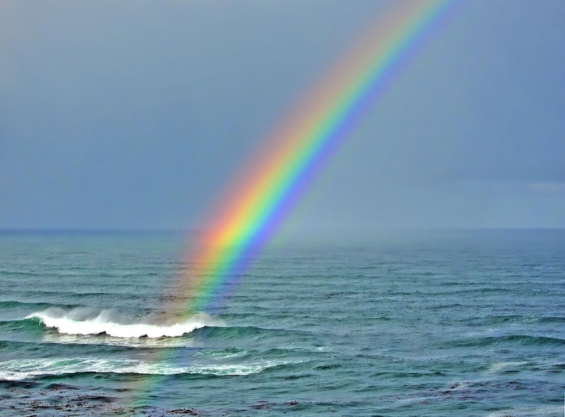World Oceans Day: Vivid rainbow over the ocean with waves and a blue sky.