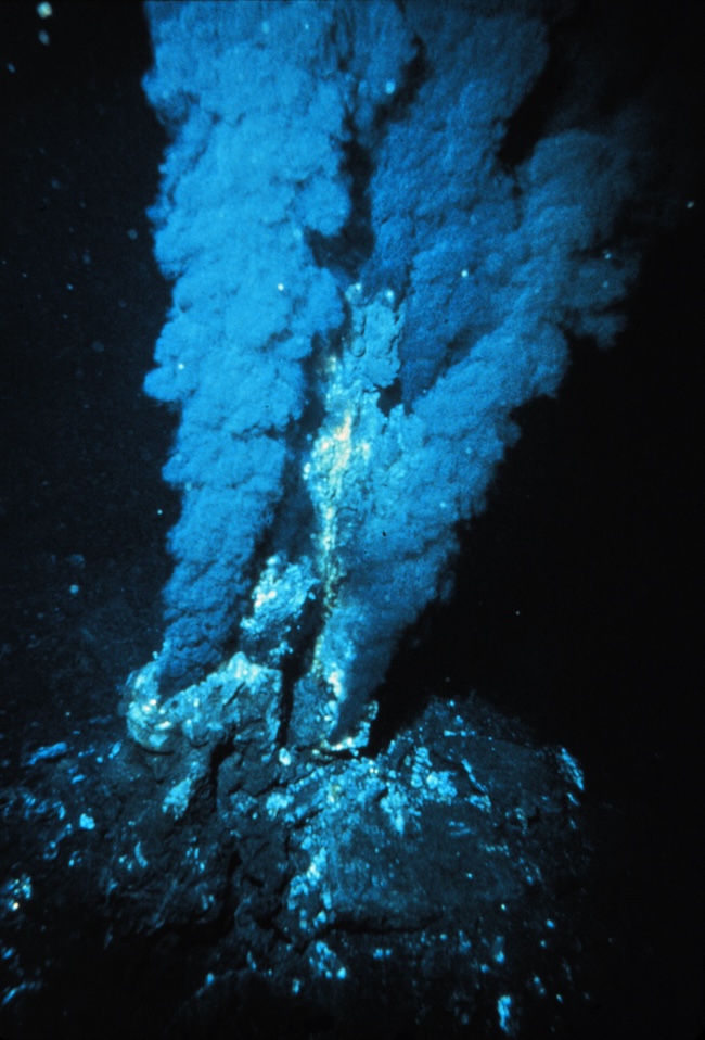 Billowing geyser-like pillars of smoke coming from holes in a rock in deep dark water.