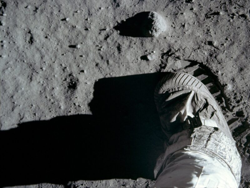 Footsteps on the moon: A view straight down onto moon boot hovering over partly visible ridged boot print stamped into dust.