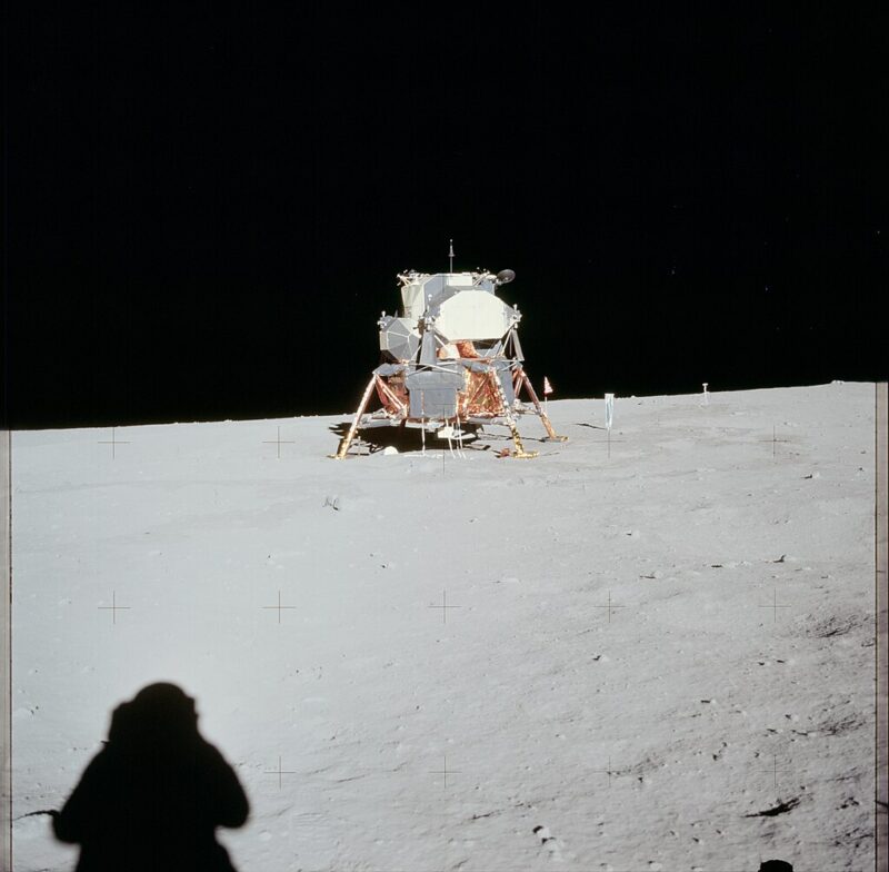 Sunlit side of boxy spacecraft on bent legs in distance, with astronaut's shadow on the ground.