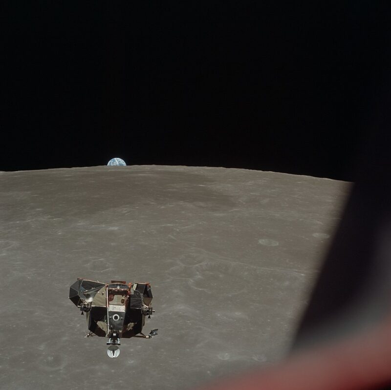 Boxy spacecraft in middle distance high above lunar surface, with Earth peeking up over the horizon.
