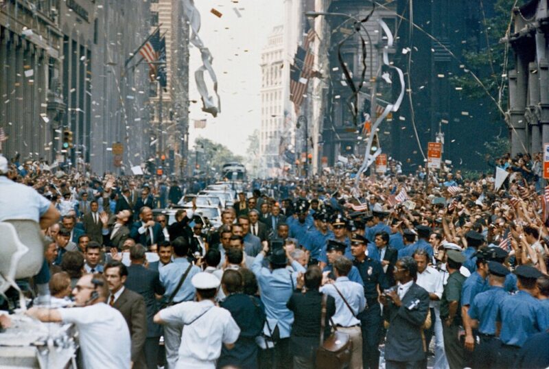 Crowds of people in street between tall buildings with air full of paper bits and streamers.