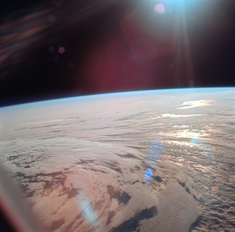 Orbital view of mostly clouded Earth with sun glinting from sea, blue along curved horizon, black sky.