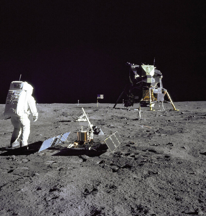 Astronaut in foreground with complicated device on the ground, lunar lander and flag in background.