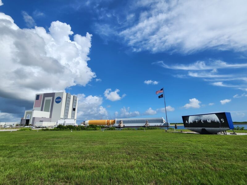 Artemis: Cloudy bright sky over green grass. Enormous building on left. Long, tubular rocket body leaving barge in center. Electric sign on right.