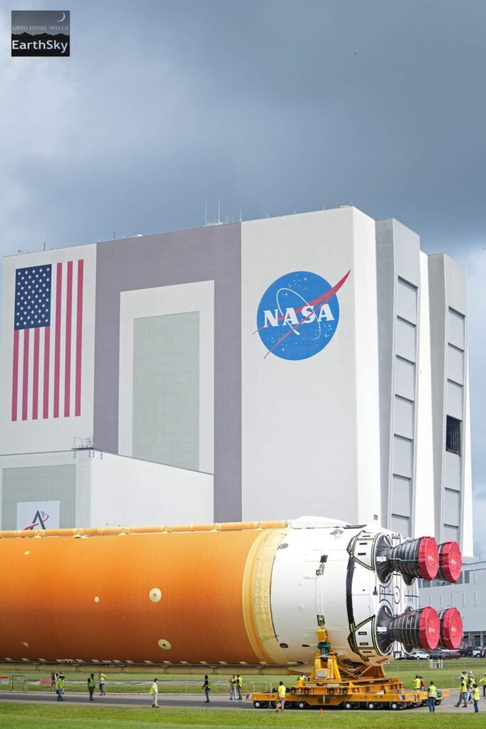 Cloudy sky over large building with American flag and NASA logo. A long rocket body lies on its side in front the building. People are gathered around it.