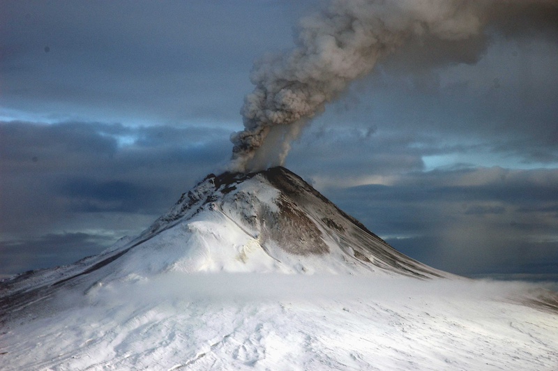 Volcano with dark smoke billowing from its peak, with snow on its slopes and dark brooding sky behind it.