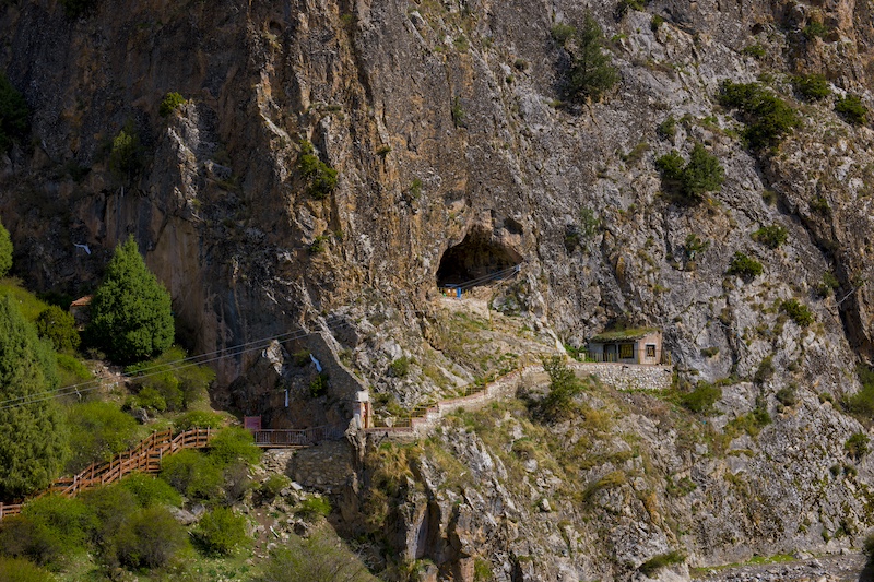 Extinct humans Denisovans: A path out to a cave and small hut in the face of a tall rocky cliff.