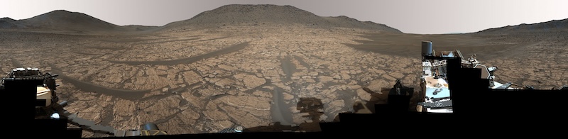 Flat cracked rocky terrain with hills in the distance and part of a robotic machine in foreground.