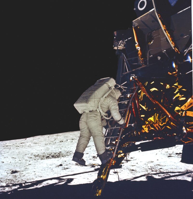 Astronaut backing down a short ladder, gold covered body of lander to right, white landscape and black sky.