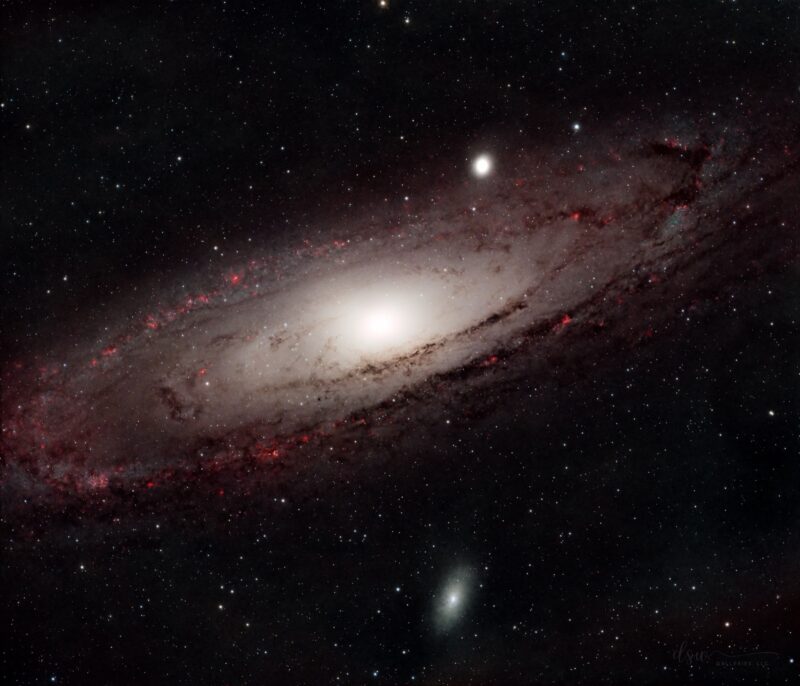 Oblique view of a large white disk-shaped nebula with bright reddish spots, dark lanes and thousands of foreground stars.