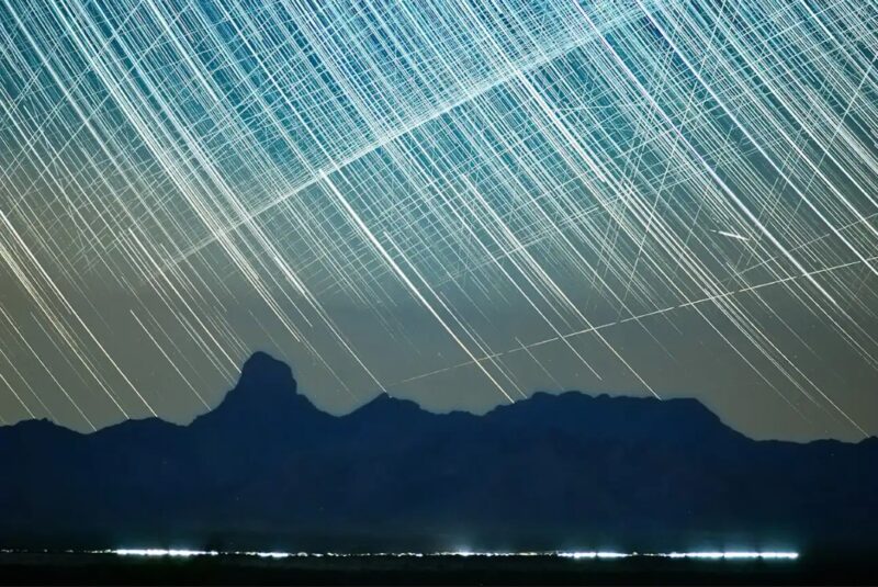 Satellite trails: City lights at the base of a dark mountain, with the sky crisscrossed with white lines.