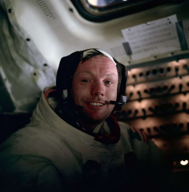 Boyish looking man in space suit with helmet off grinning at the camera.