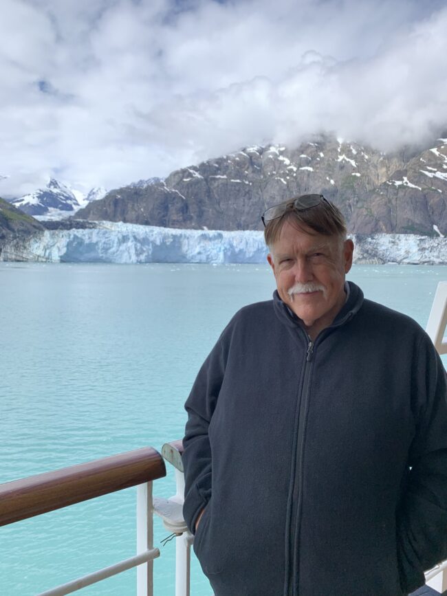 A older man wearing a dark-colored zipped-up jacket, standing on what looks like a boat, in what looks like Antarctica.