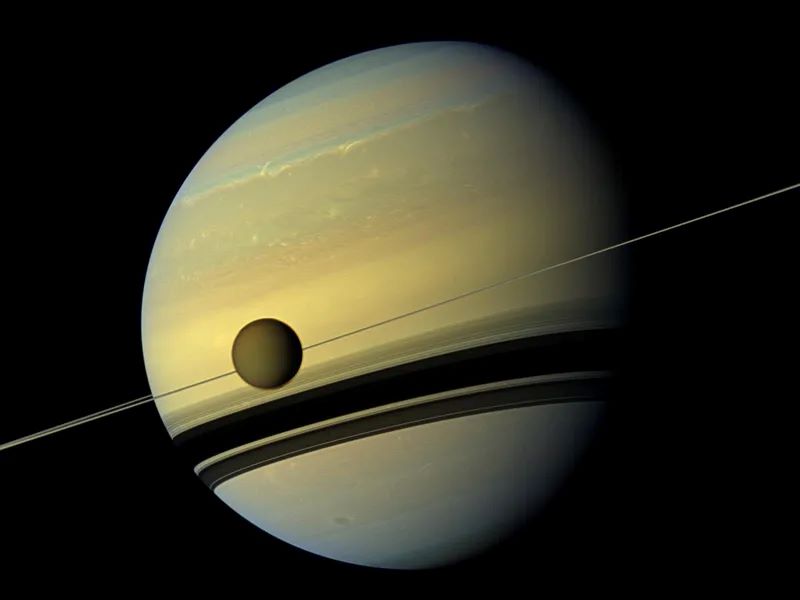 Golden globe of Saturn with edge-on rings and dark moon, and wide shadows of rings striping the planet.