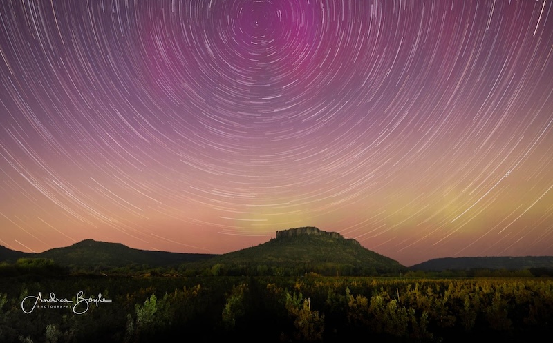 Semicircle of thin streaks of light from star trails in a pink and yellow sky with hills in the foreground.