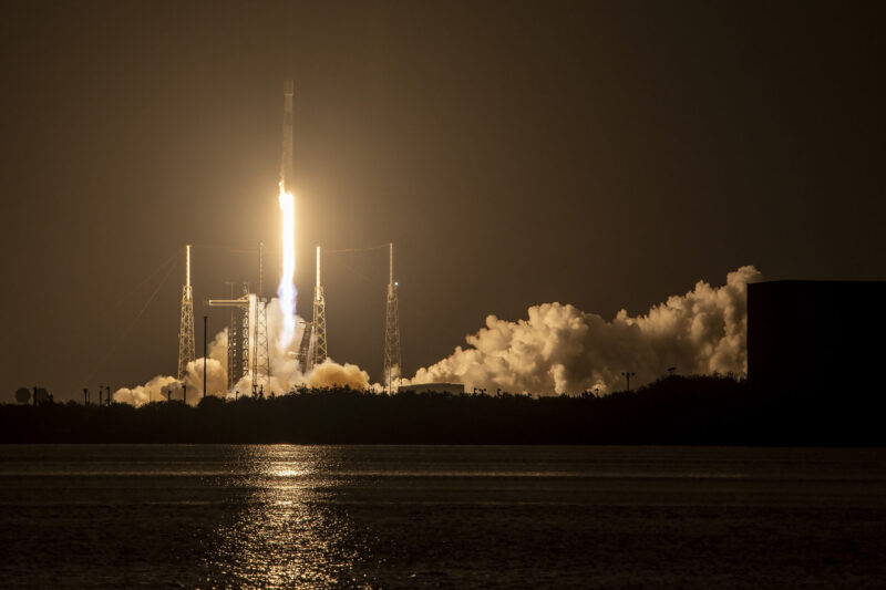 A rocket on a pillar of fire launching at night with reflection in the water.