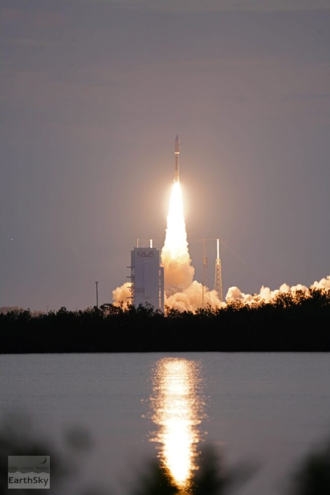 From a distance across water, a rocket with flames below launches upward.