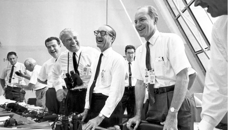 Eight happy, laughing men in white shirts with dark ties standing by control panels.