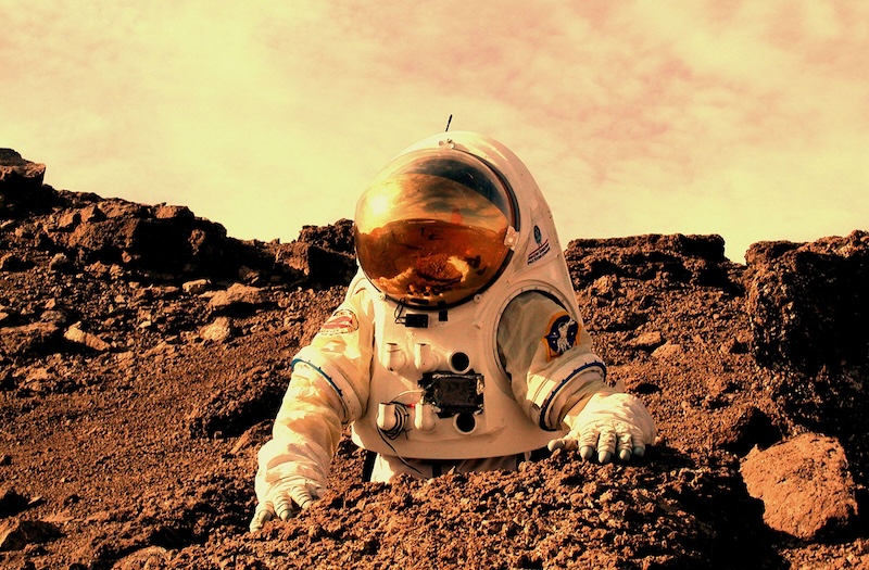 Astronaut in bulky white suit examining rocks in reddish terrain with dusty sky above.