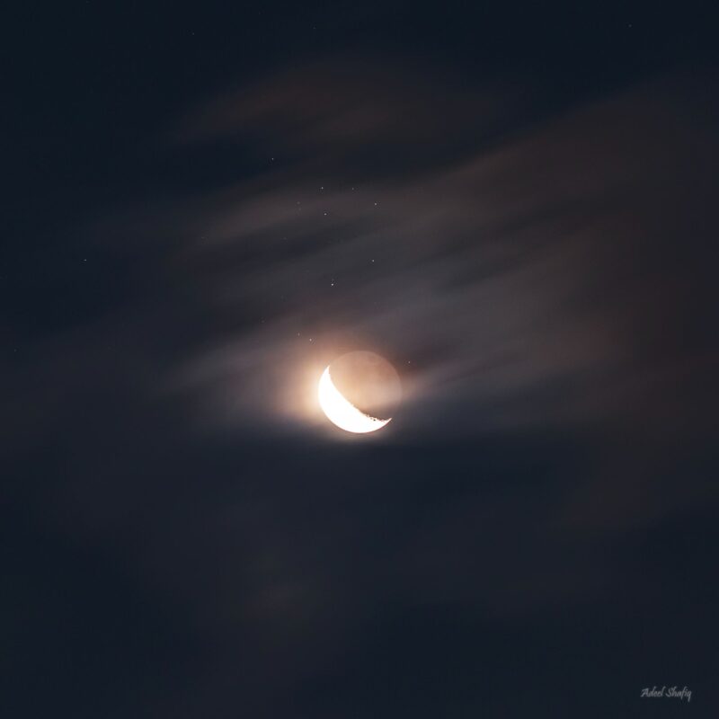 Left side of the moon shining. There are some thin clouds at the top right of the image and several dots above the moon.