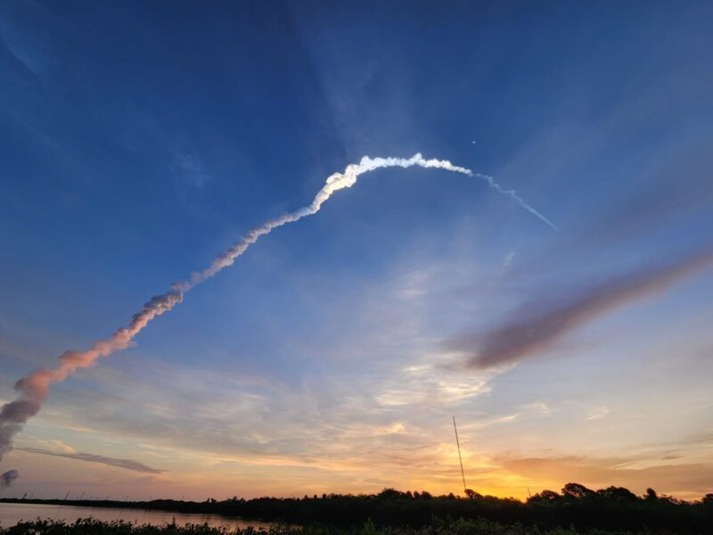 A horizon glowing with the coming sunrise and a plume of smoke heading upward into blue skies, turning white higher in the air.