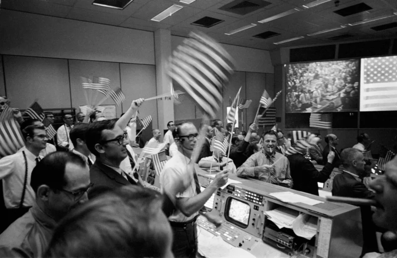 Roomful of exhilarated men standing and waving American flags, control panels visible.