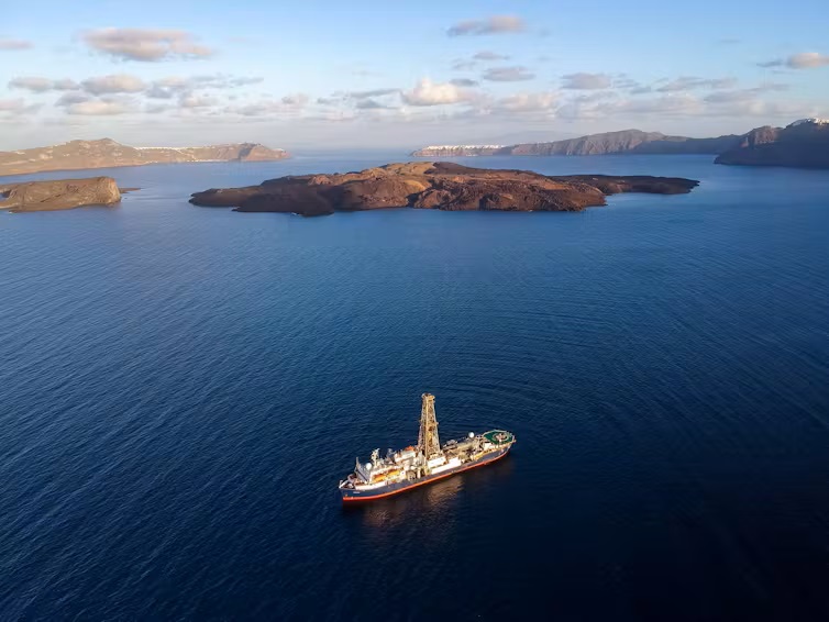 Volcanic eruption: Aerial view of a ship with a tall drill rig on it and small islands poking up all around in a blue sea.