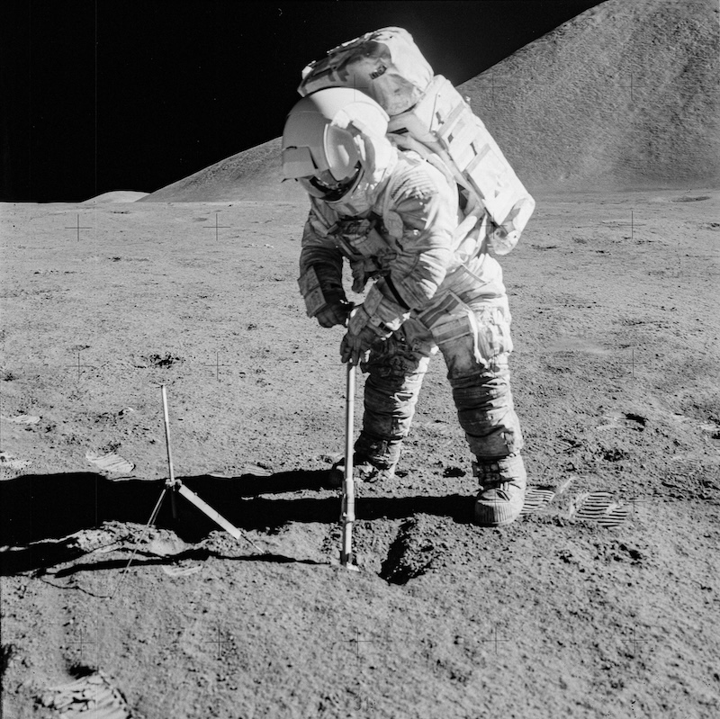 Astronaut in bulky white spacesuit standing on the moon's desolate terrain under a black sky.