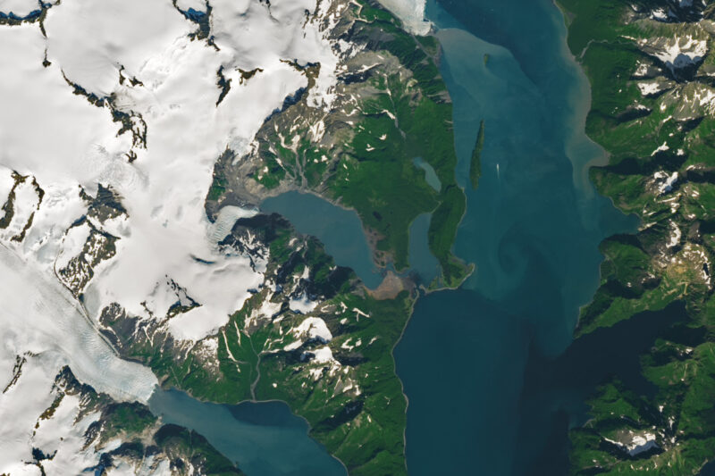 Satellite view of green land with brown swirls and blue water on either side and ice caps on the left.