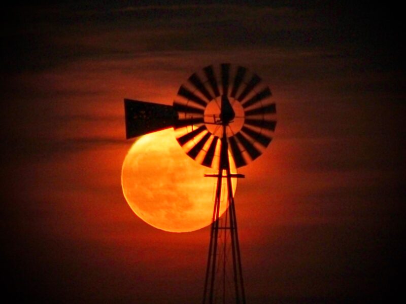 Orange, big, bright moon behind a windmill. Some haze surrounds the view.