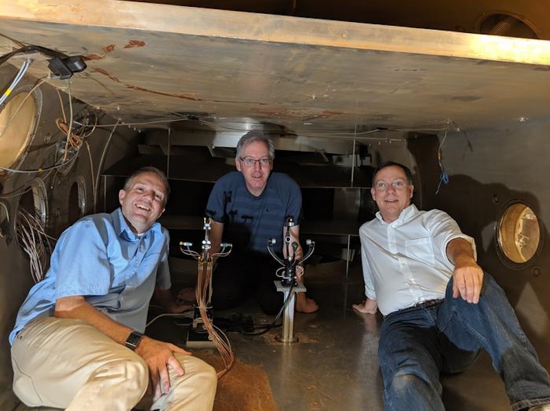 Winds on Mars: 3 men sitting inside a small enclosed space, with 2 small mechanical instruments between them.