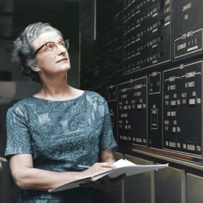 Middle-aged woman in a blue dress holding a notebook and looking up at readouts from a giant computer in the 1960s.