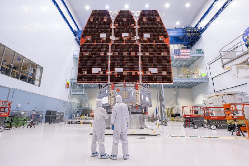 Two people in white clean suits inside a large building looking up at three solar panels.