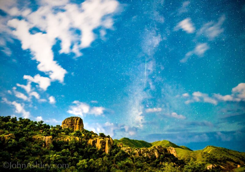 Light blue sky with some white clouds. There is a mountain with trees in the foreground. A band of stars with a long streak in the middle.