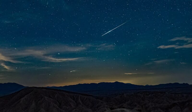 Blue, starry sky with some light clouds and a orangish horizon. There are some mountains below.