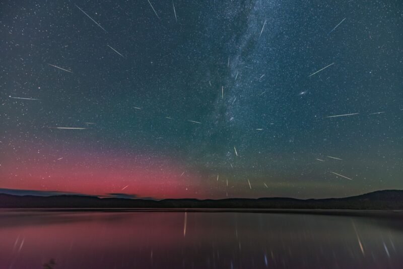 Starry blue sky with some red in the horizon, to the left side, and green on the rigth side. Many white streaks cross the sky and are reflected in the water.