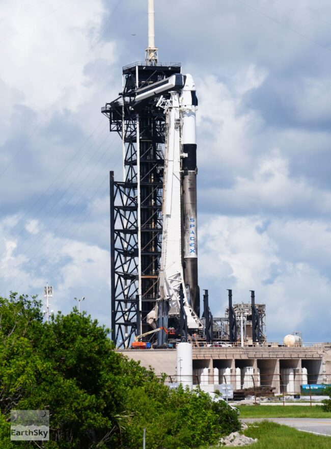 Daylight sky covered on clouds. There is a white and black rocket next to a big structure that is holding the top of the rocket.