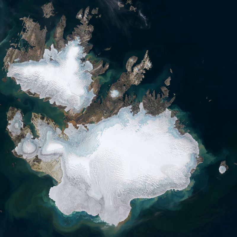 Svalbard is melting: Satellite view of islands in a dark sea, with rocky edges, nearly covered with ice.