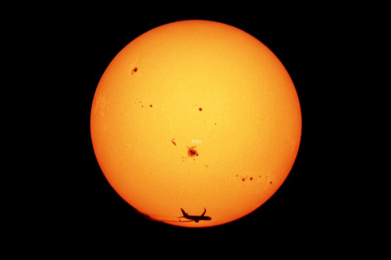 The sun, seen as a large yellowish sphere with small dark spots and an airplane flying through.