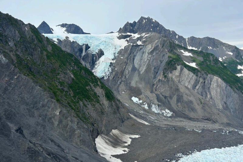 Rugged gray mountains with a glacier at top and a long scar from the glacier to the sea below.