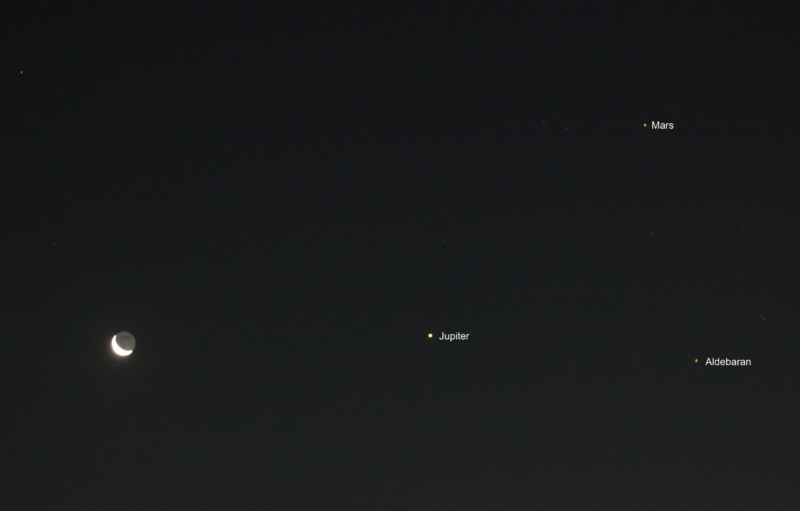 Dark sky with the moon on the left side. A dot for Jupiter to its right side. Two reddish dots to the right of Jupiter, above and below, creating a triangle.