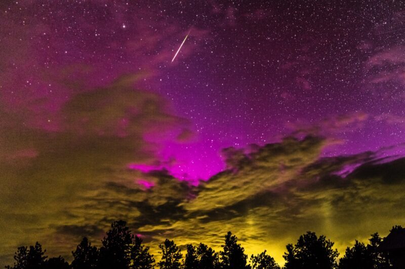 Very bright pink sky with strong yellow clouds below. There are many trees at the bottom of the image. A bright streak crosses the sky.