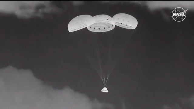 Space capsule in midair tied to the bottom of 3 parachutes.