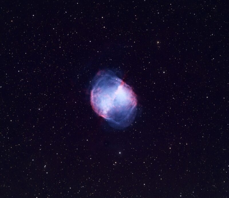 Dumbbell-shaped cloud of reddish and blue nebulosity over a background of distant stars.