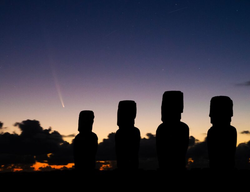 Silhouettes of heads on Easter Island with sunrise colors and a comet at left.