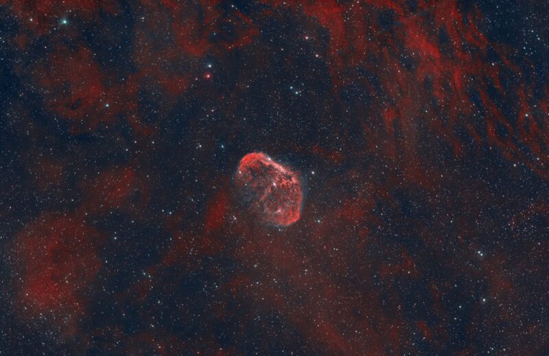 A small and bright, reddish cocoon of gas overlaid with prolific red clouds and numerous stars.