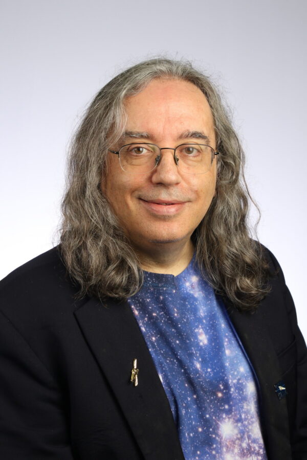 Space Race: Man with long gray hair smiles in a portrait. He is wearing a star-studded shirt and a dark blazer.